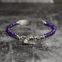 A beaded bracelet with faceted purple amethyst gemstones and Argentium Sterling Silver bar and extension chain, including a small charm, displayed on a textured gray surface