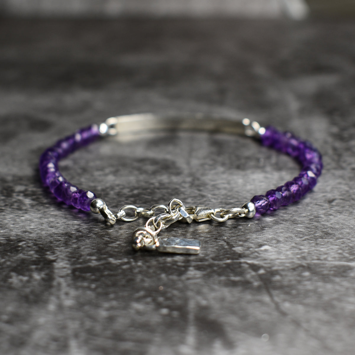 A beaded bracelet with faceted purple amethyst gemstones and Argentium Sterling Silver bar and extension chain, including a small charm, displayed on a textured gray surface