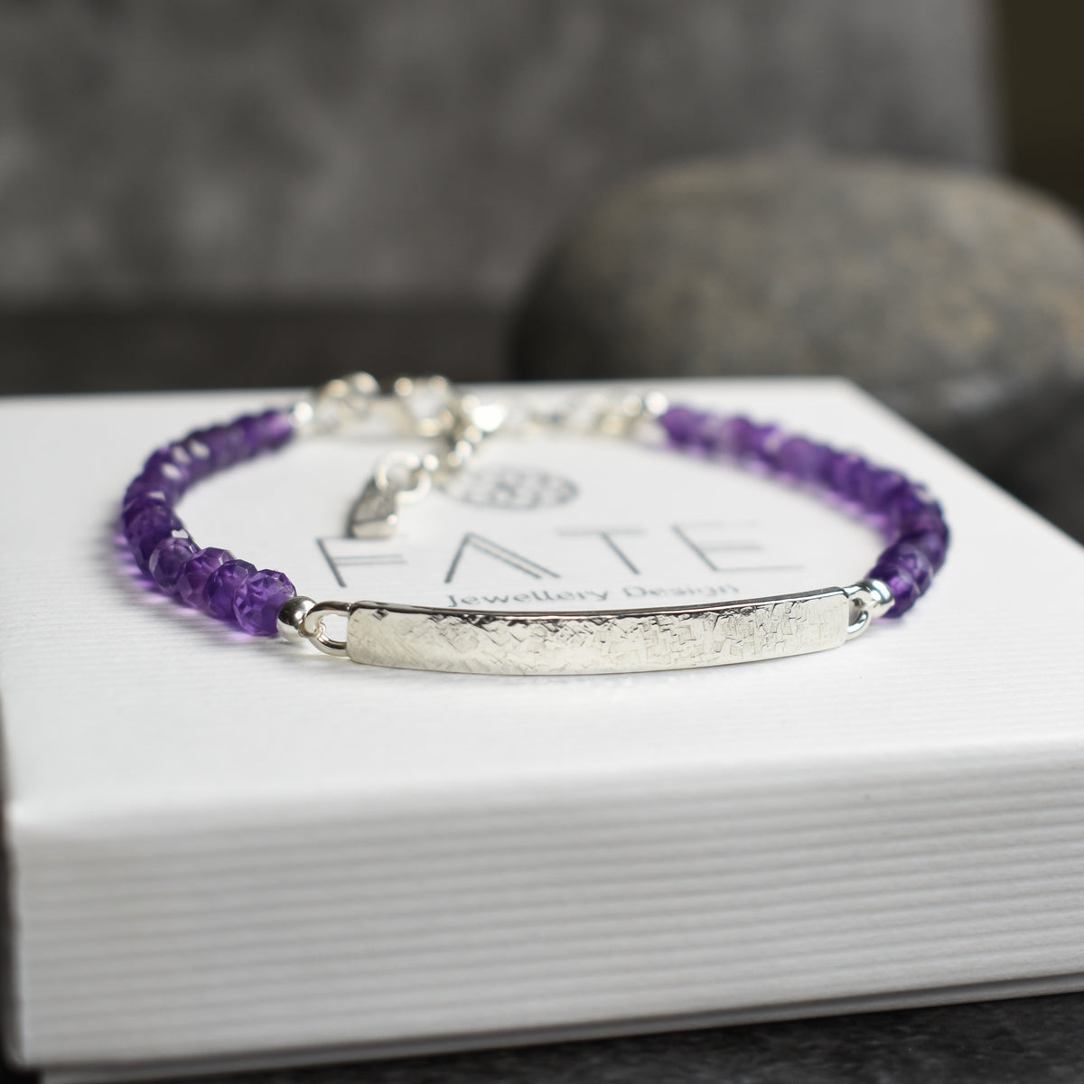 A beaded purple and silver bracelet resting on a white FATE jewelry gift box, with a blurred gray background.