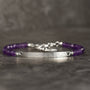 A Amethyst beaded bracelet with faceted purple beads, Sterling silver spacer beads, and a hammered silver ID bar, resting on a dark gray textured surface.
