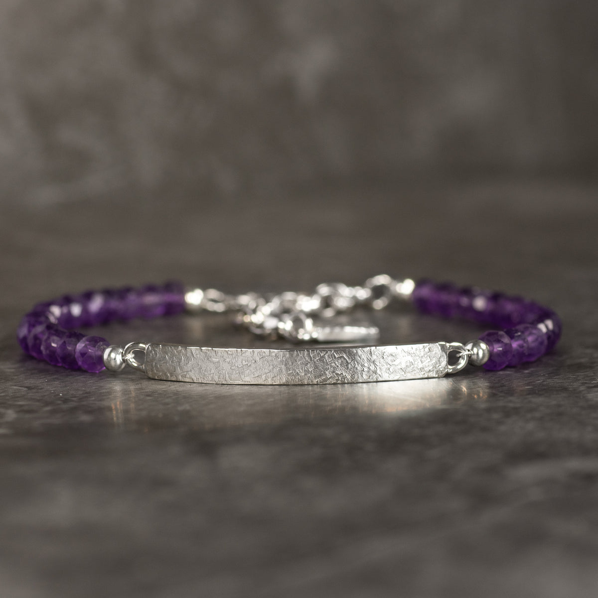 A Amethyst beaded bracelet with faceted purple beads, Sterling silver spacer beads, and a hammered silver ID bar, resting on a dark gray textured surface.