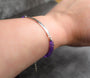 A close-up of a person's wrist wearing a silver-toned amethyst gemstone bracelet with purple faceted bead accents on both ends, set against a dark gray textured background.
