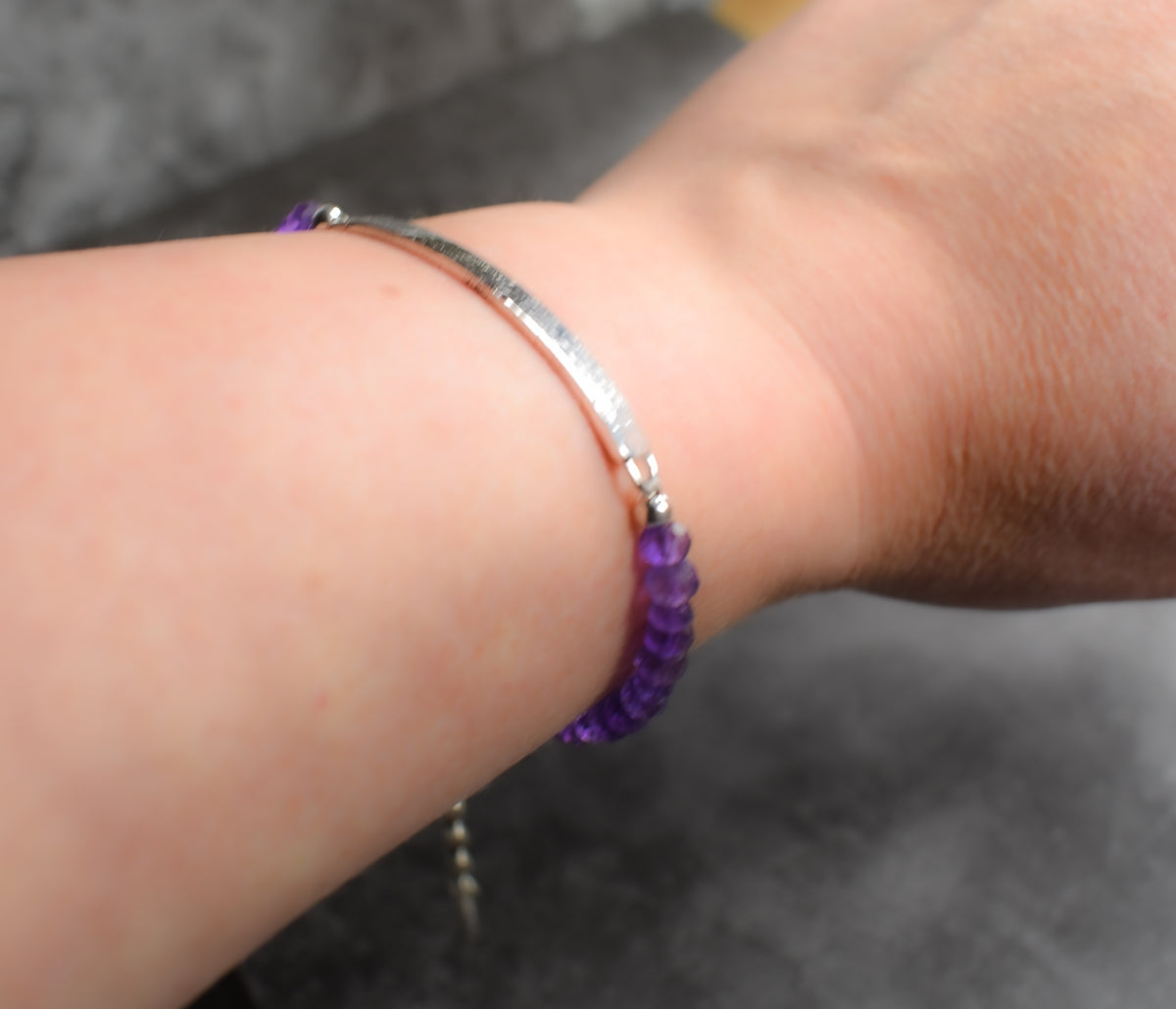 A close-up of a person's wrist wearing a silver-toned amethyst gemstone bracelet with purple faceted bead accents on both ends, set against a dark gray textured background.