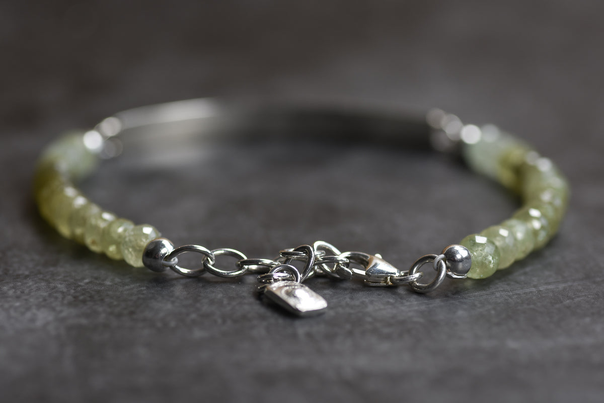 A beaded bracelet with pale yellow-green faceted beads and silver-tone chain detailing, clasp, and small hanging tag, photographed on a dark gray textured surface.