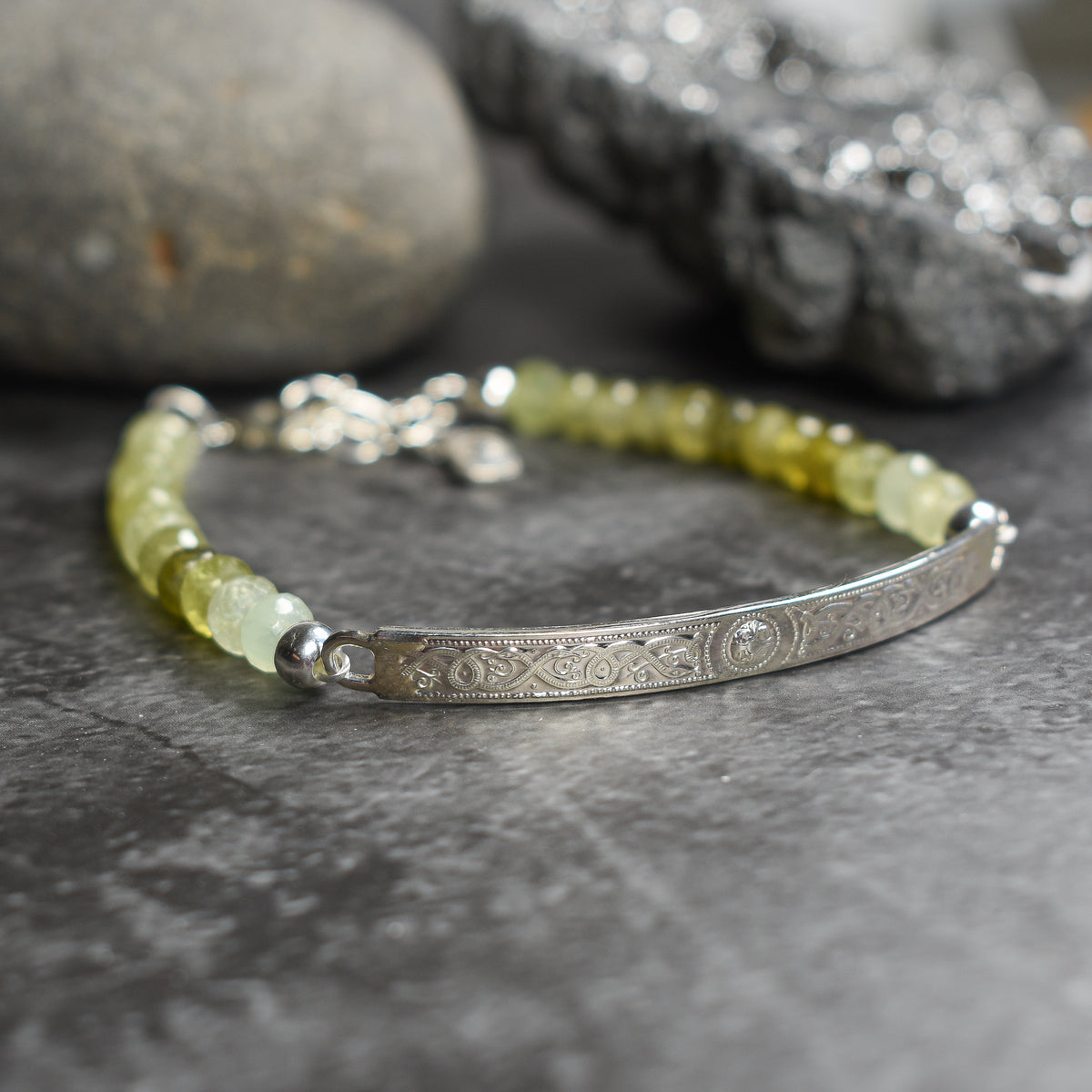 A beaded bracelet with pale yellow-green translucent beads and an engraved silver bar, photographed on a dark textured surface with blurred stones in the background.