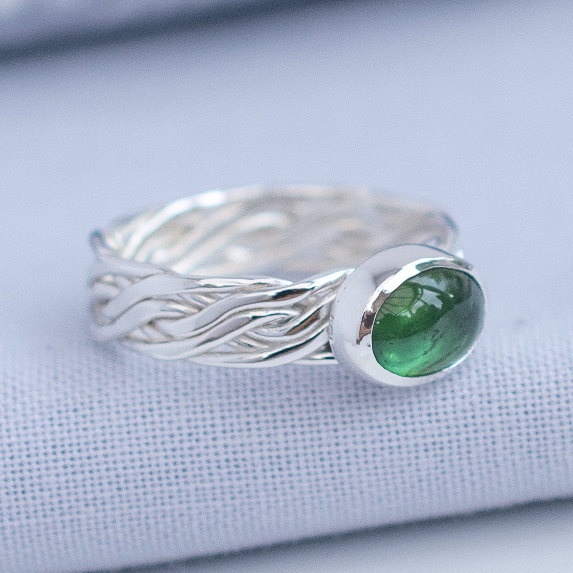 A silver ring with a braided, wave-like band and a bezel-set oval green gemstone, resting on a textured light blue fabric surface.