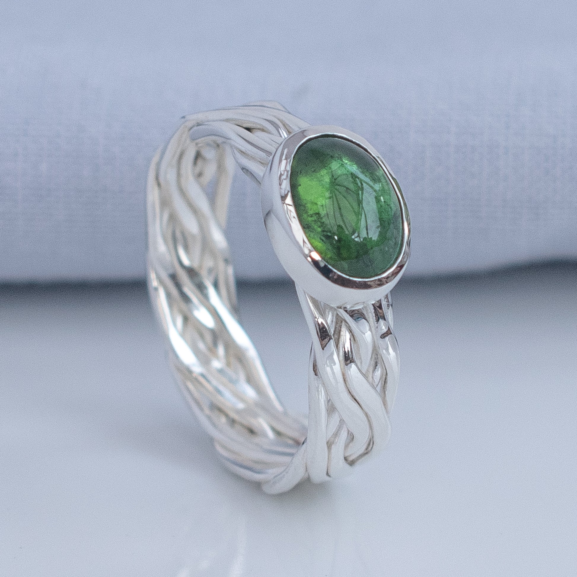 A silver ring with a twisted, braided band and an oval green gemstone, shown on a light gray surface with a soft gray fabric background.