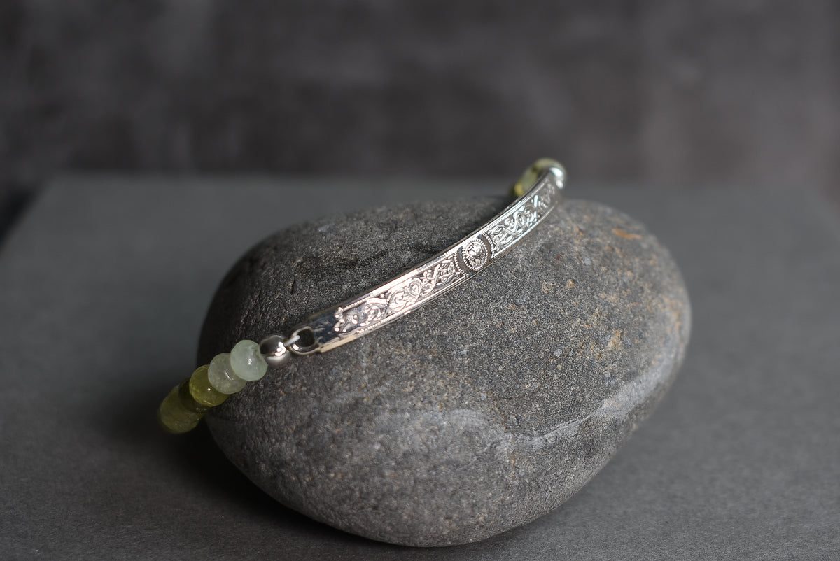 A sterling silver celtic engraved bar bracelet with 5mm faceted pale green to dark green gemstone beads, displayed on a smooth dark gray stone against a dark gray background.