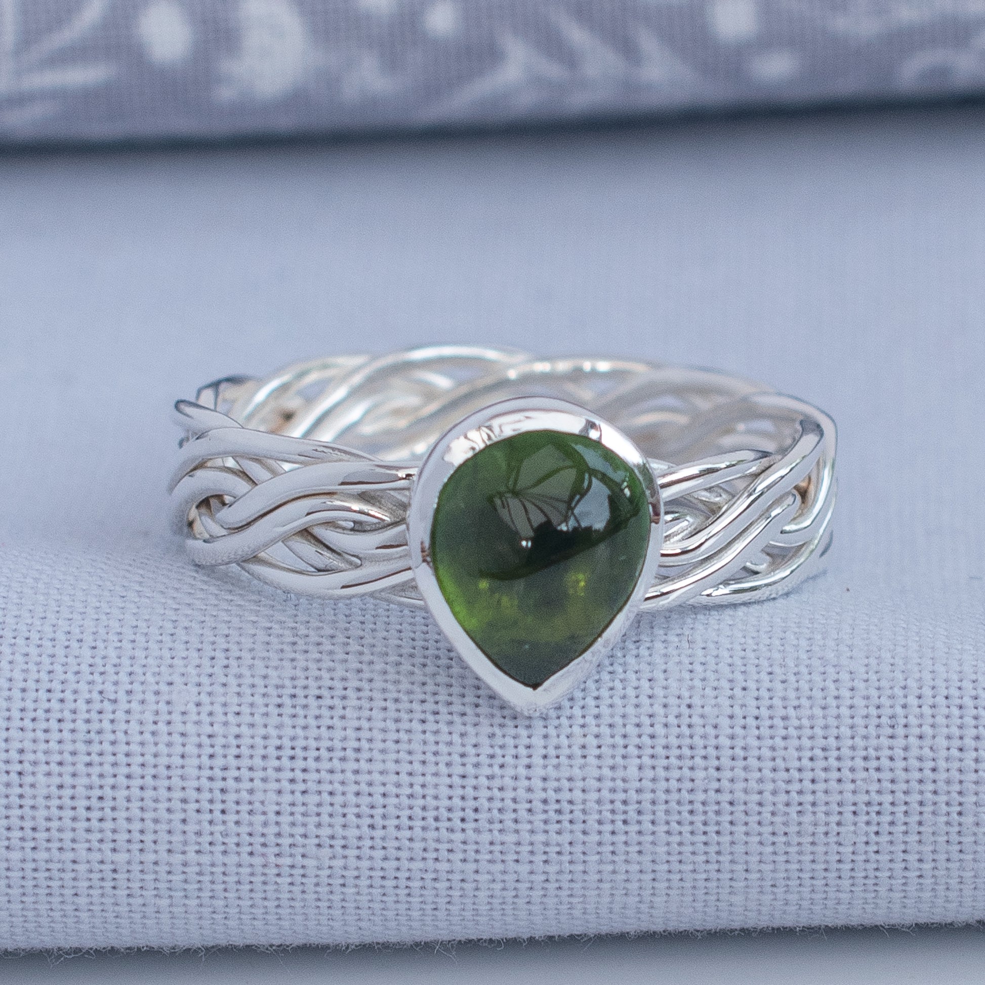 A sterling silver ring with a twisted, braided band and a teardrop-shaped green gemstone, displayed on a textured light blue fabric surface.