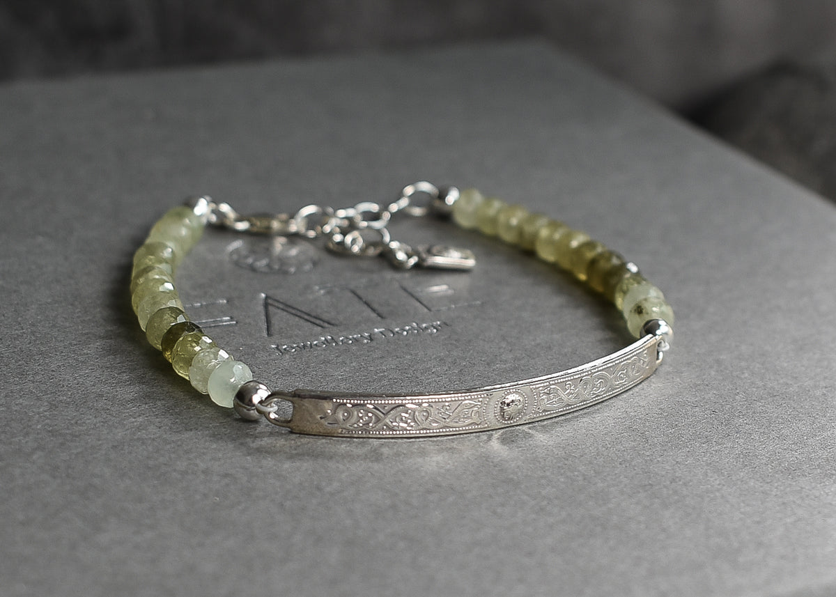 A gemstone neaded bar bracelet featuring a textured sterling silver ID style Celtic bar and a beaded strand of pale yellow-green gemstone beads, resting on a gray textured surface.